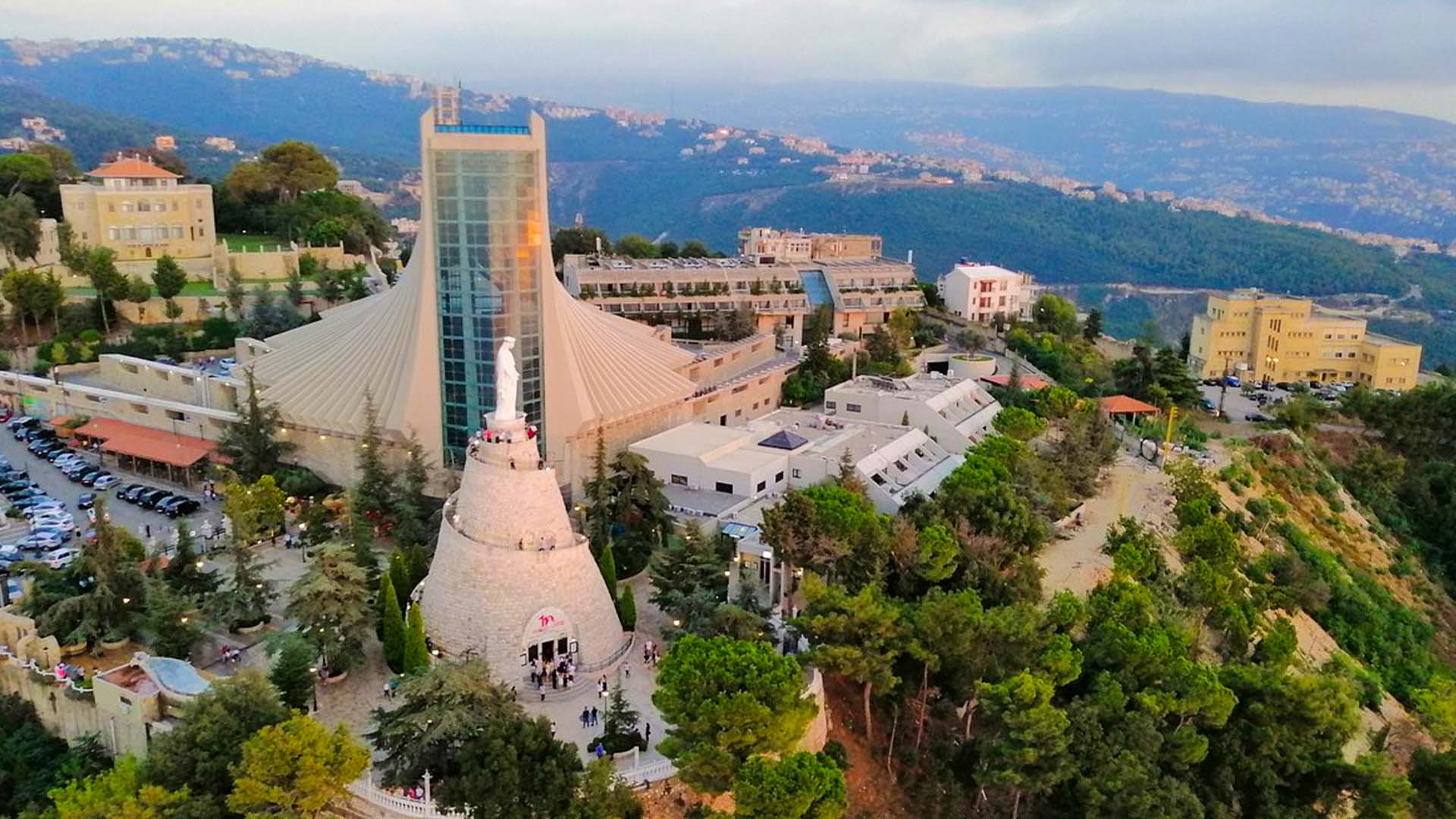 A panoramic photograph immortalizes the regal statue of Our Lady of Lebanon, situated in Harissa, embraced by verdant greenery and framed by majestic mountains.