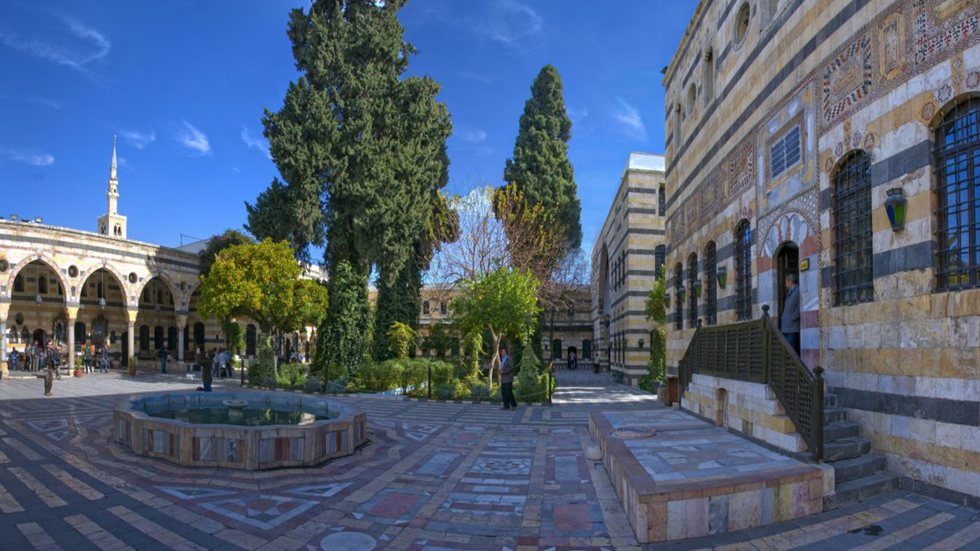 A photograph showcases the expansive courtyard of Al-Azem Palace in Damascus, adorned with towering trees in the background.