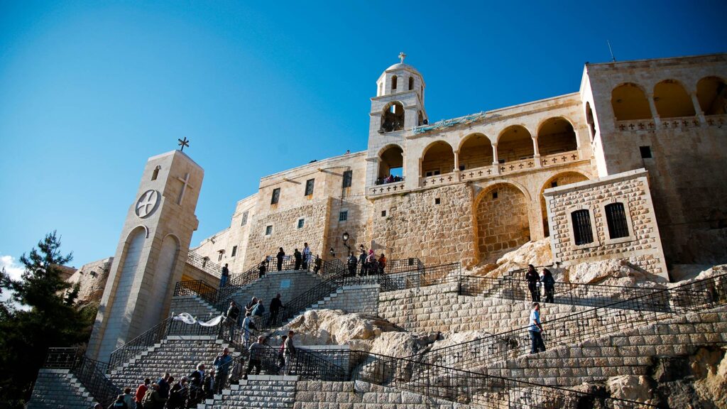 A panoramic photograph showcases the majestic Monastery of Our Lady of Sednaya, while in the background, visitors can be seen ascending the stairway, adding a sense of scale to the scene.