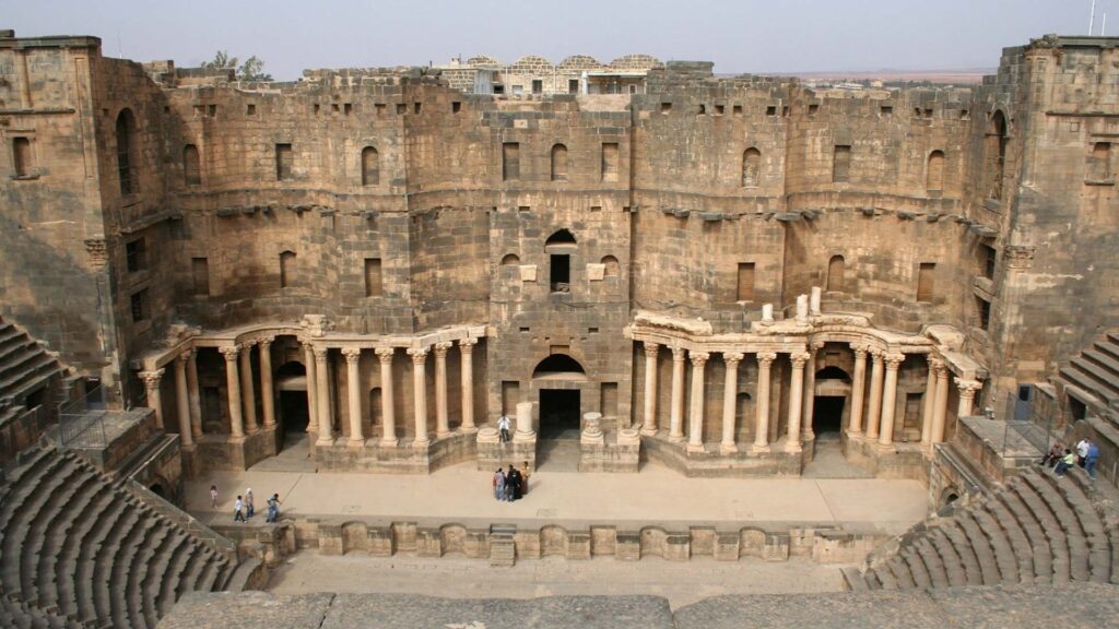 A captivating scene emerges as the Roman Amphitheatre of Bosra comes into view, its grand courtyard adorned with a scattering of tourists, creating a picturesque tableau.