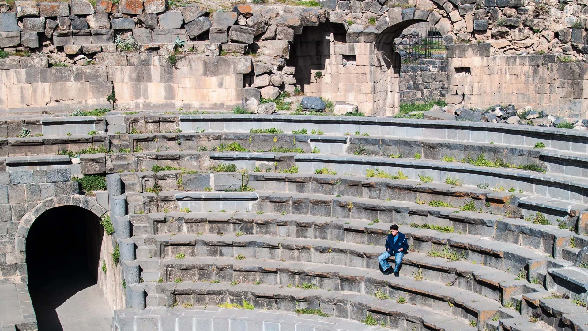 The panoramic photograph immortalizes the Roman theater of Shahba, while a local resident relaxes on the stone seats, adding a touch of contemporary life to the historical setting.