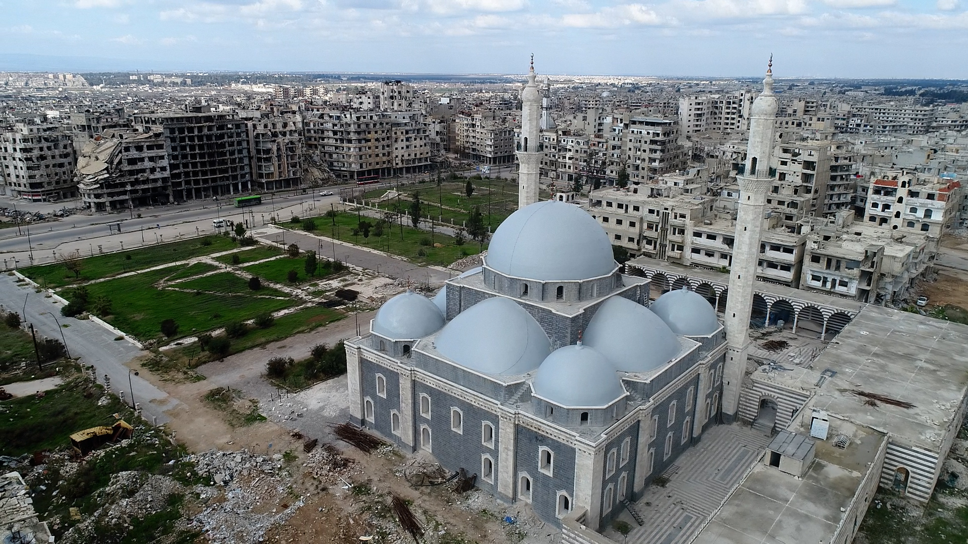 Taken from above, a photograph captures Khalid Ibn al-Walid Mosque of Homs, with a visibly damaged building serving as a poignant backdrop, creating a compelling visual contrast.