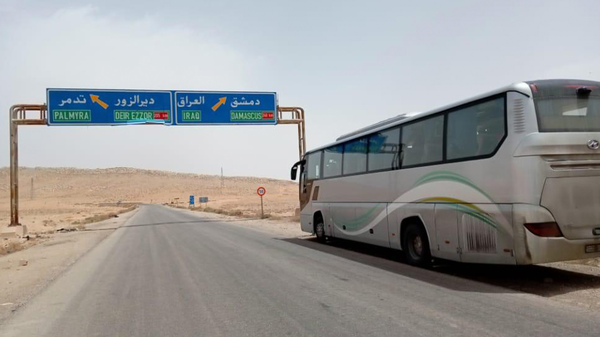 In the photograph, a tourist bus can be seen passing beneath a blue signboard that displays the destinations of Palmyra, Deir EzZor, Iraq and Damascus.