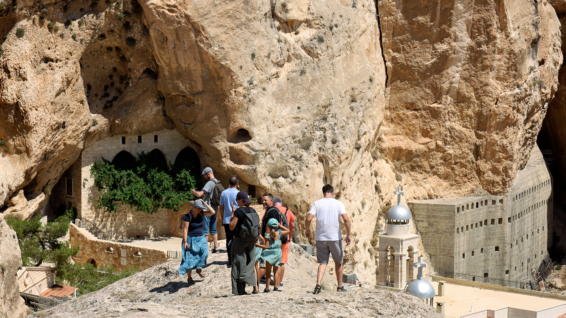 A photograph captures a group of tourists strolling towards the churches of Maalula, with a massive rock looming in the background.