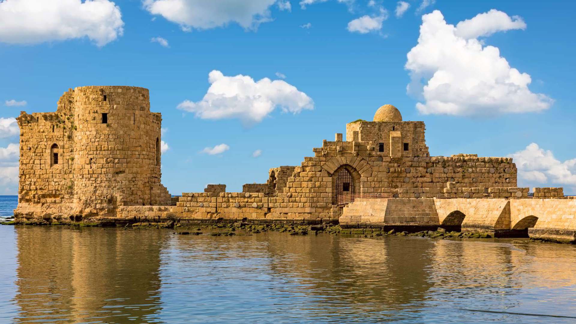 A photograph captures the Sea Castle of Sidon, perched on a small island in the midst of the Mediterranean Sea and linked to the mainland by a slender causeway.