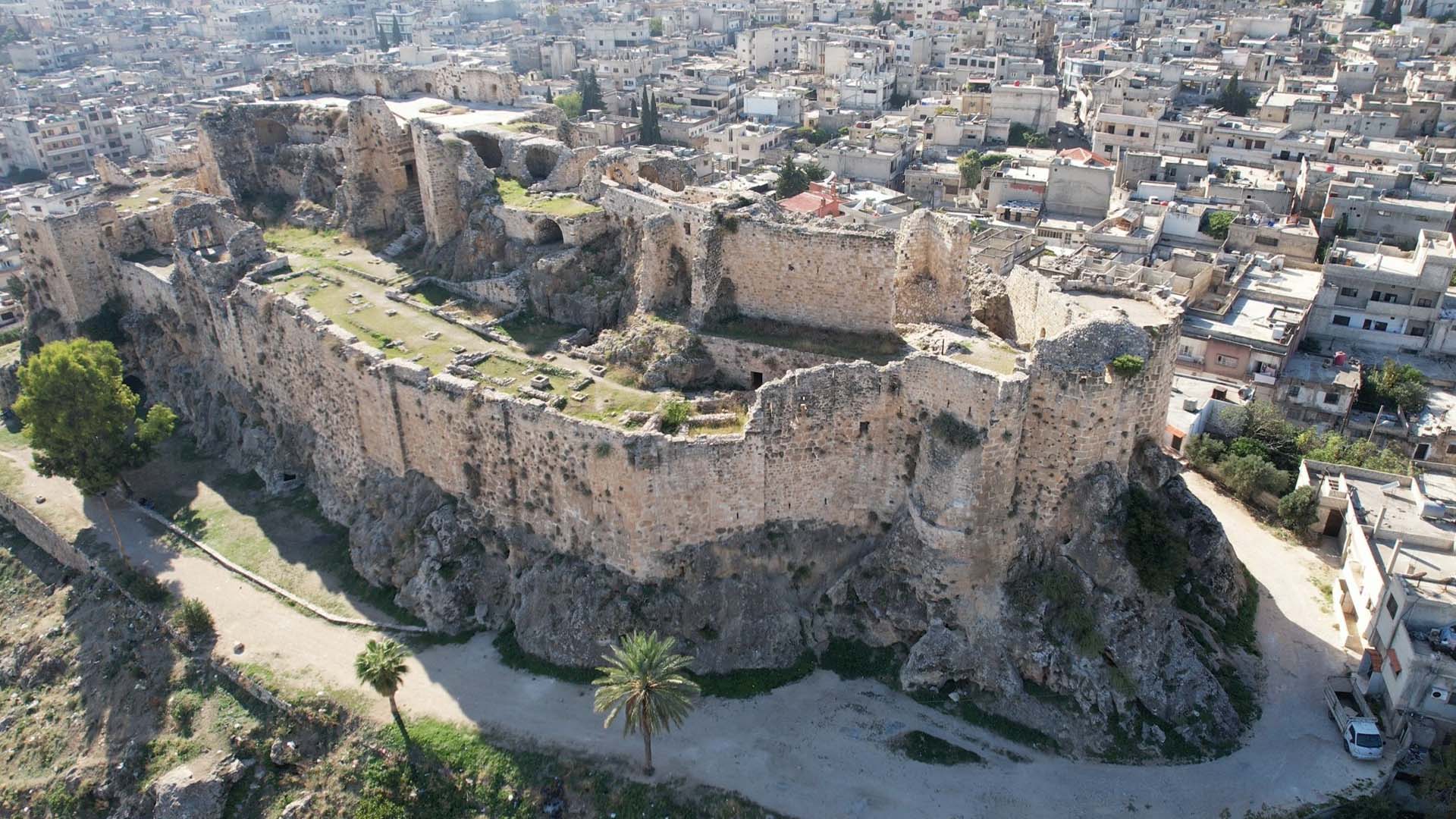 From an aerial perspective, a photograph showcases the magnificent Masyaf Castle, strategically situated atop a hill, while houses are scattered in the background, creating a picturesque scene.
