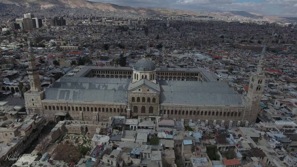 An aerial photograph captures the Umayyad Mosque, the iconic landmark of Damascus, gracefully standing in the background amidst a scattering of houses, with Mount Qassioun majestically looming in the distance.
