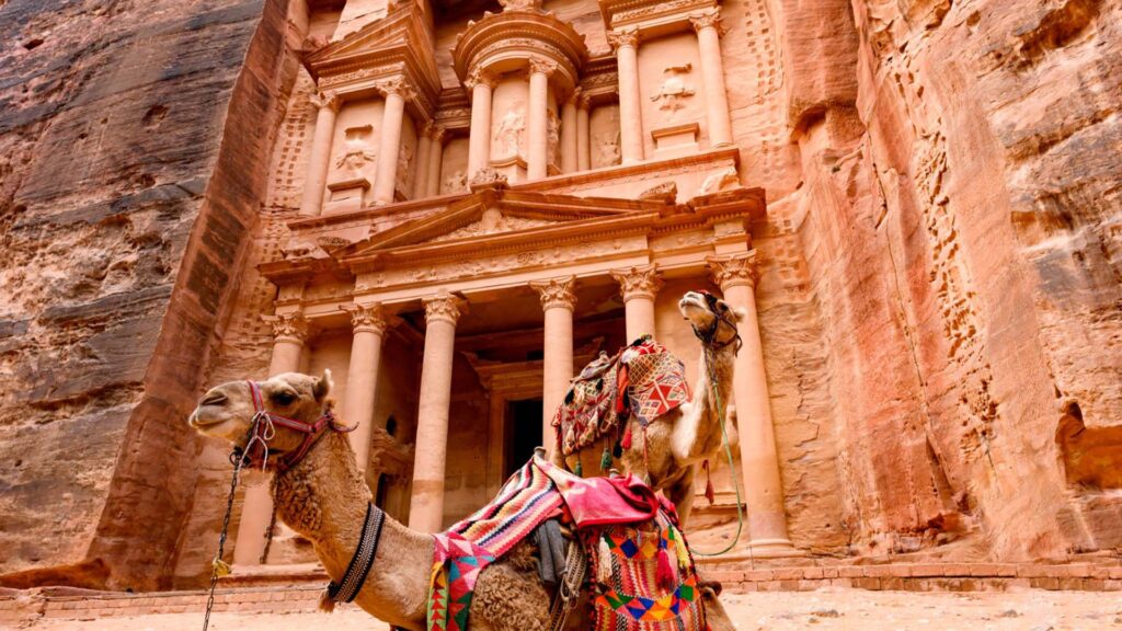 A photograph captures the timeless grandeur of Petra's rock-cut architecture, where two camels command attention in the foreground.