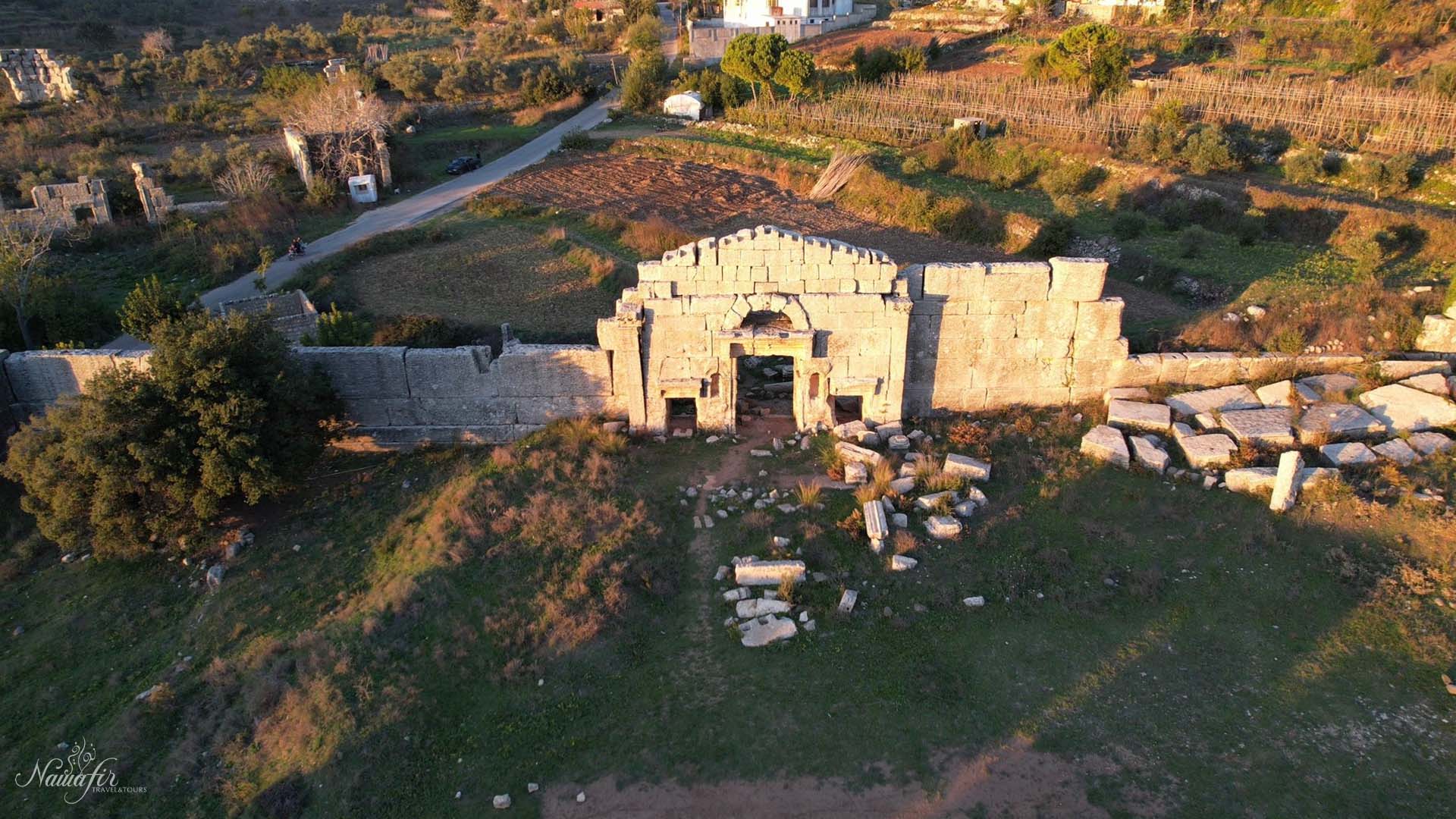 From an aerial viewpoint, a photograph captures the entrance of Hosn Suleiman, embraced by lush trees and scattered ruins on the ground, creating a captivating composition.