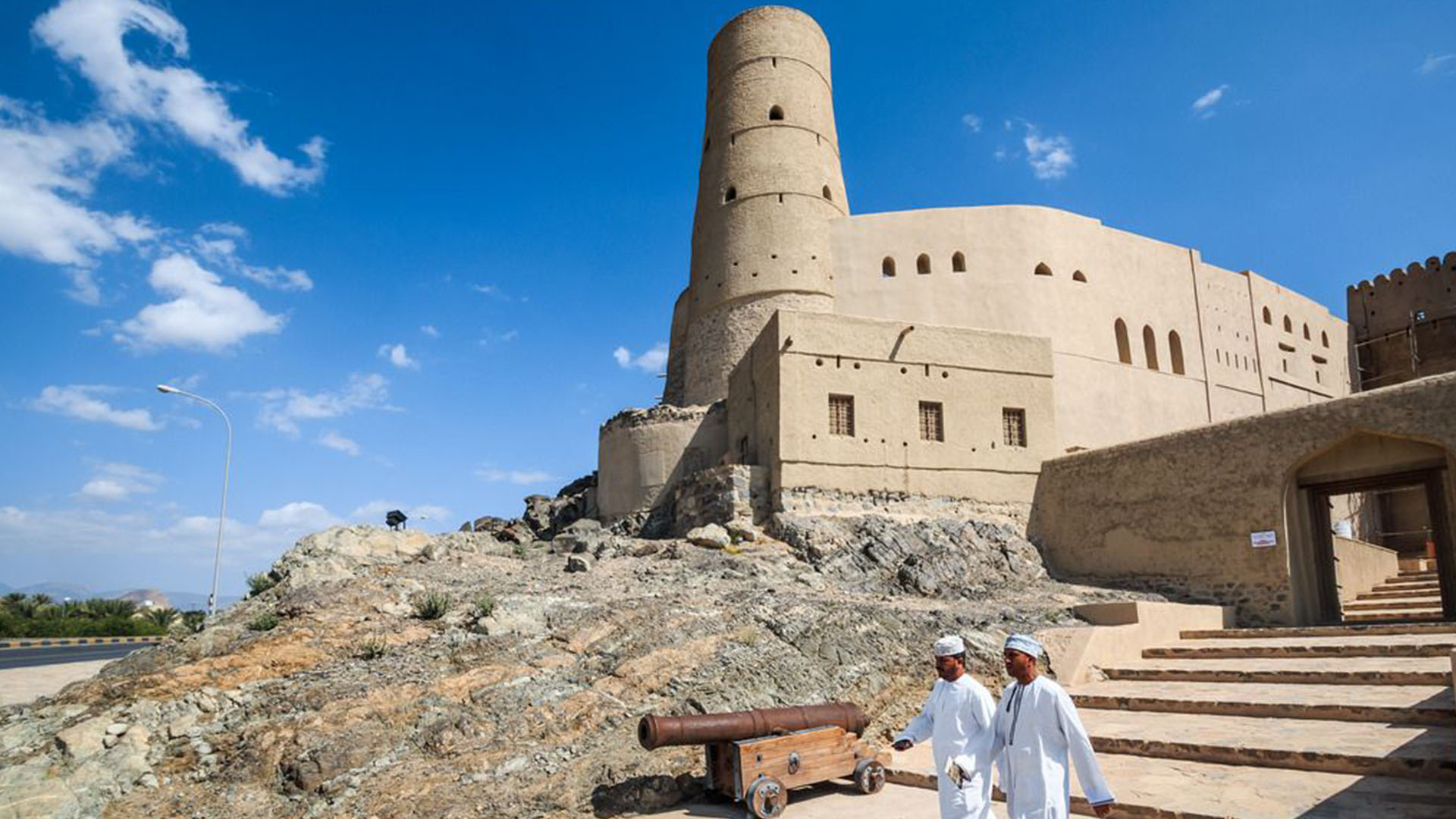 A panoramic photograph immortalizes the commanding presence of Bahla Fort, as it dominates the skyline of the town, while locals descend the stairway, adding a sense of movement and vitality to the scene.