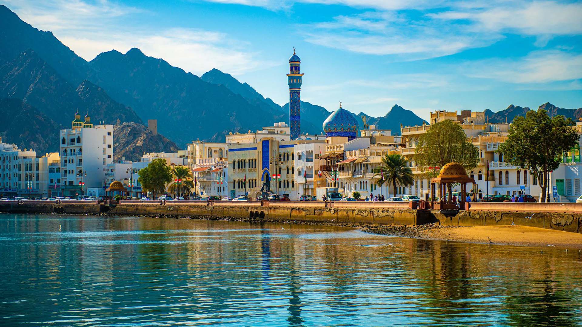 A panoramic photograph captures the stunning coastal front of Muscat, where a prominent mosque takes center stage amidst a backdrop of buildings, while a majestic mountain looms in the distance.