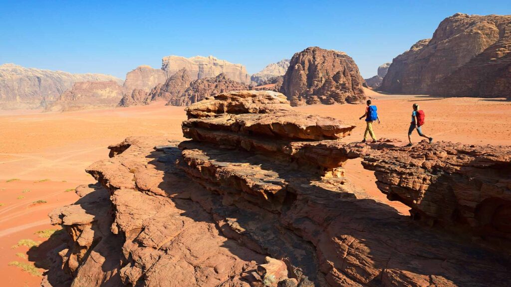 A panoramic photograph captures the timeless adventure of tourists scaling the rocks of Wadi Rum, with mighty mountains standing tall in the background.